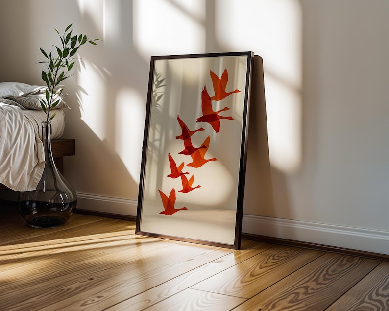 May include: A framed art print featuring a flock of orange birds in flight against a cream background. The artwork is displayed in a dark brown frame and leans against a white wall. The room has hardwood floors and a glass vase with greenery.