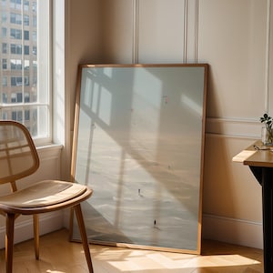 May include: A framed landscape art print leans against a wall next to a wooden chair with a woven seat. The artwork depicts a beach scene with figures. A small table with a plant and a notebook is in the corner.