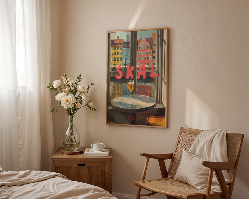 May include: A framed art print featuring a colorful cityscape with the word "SKAL" in red letters. A glass of beer sits on a table in front of a window. The print hangs above a wooden nightstand with a vase of white flowers.