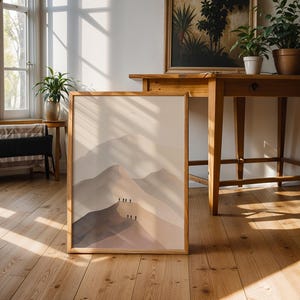 May include: A framed art print featuring a minimalist mountain landscape in shades of beige and brown. The artwork is displayed on a wooden floor, leaning against a wall. The scene includes wooden furniture and potted plants, creating a warm, inviting atmosphere.