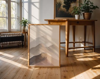May include: A framed art print featuring a minimalist mountain landscape in shades of beige and brown. The artwork is displayed on a wooden floor, leaning against a wall. The scene includes wooden furniture and potted plants, creating a warm, inviting atmosphere.