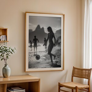 May include: Black and white framed photograph of people playing soccer on a beach. The image features a woman kicking a soccer ball, with other figures in the background. The photograph is displayed on a beige wall.