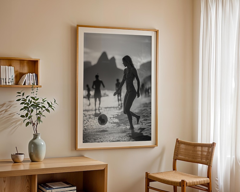 May include: Black and white framed photograph of people playing soccer on a beach. The image features a woman kicking a soccer ball, with other figures in the background. The photograph is displayed on a beige wall.