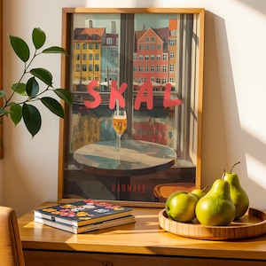 May include: A framed print with the word "SKAL" in red letters, depicting a cityscape with colorful buildings and a glass of beer. A wooden tray holds green pears. A stack of notebooks with floral designs sits on a wooden desk.