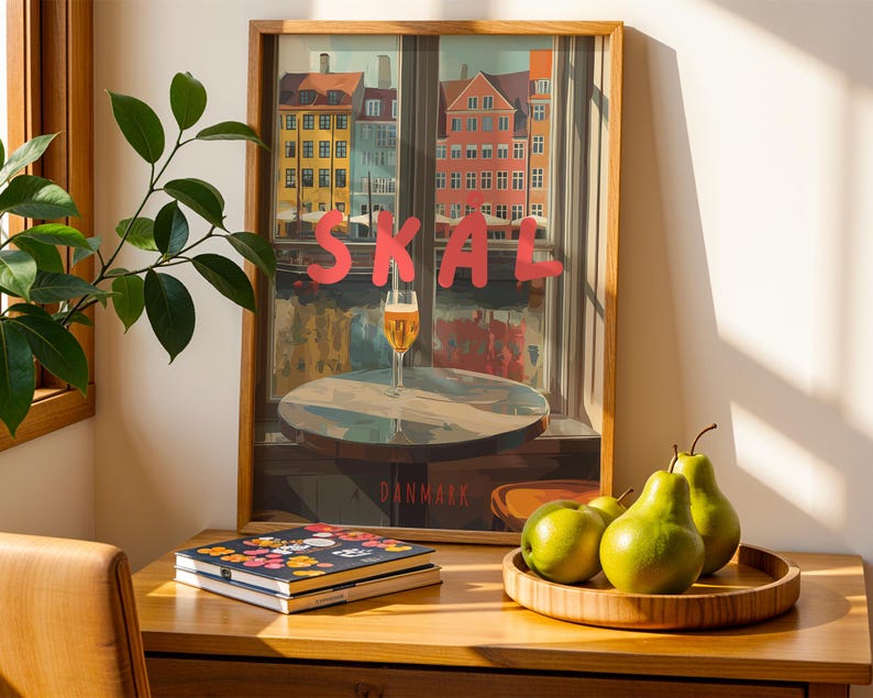 May include: A framed print with the word "SKAL" in red letters, depicting a cityscape with colorful buildings and a glass of beer. A wooden tray holds green pears. A stack of notebooks with floral designs sits on a wooden desk.