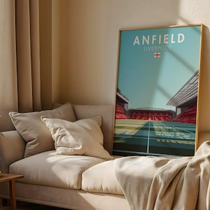 Puede incluir: Un cuadro enmarcado con una ilustración estilizada del estadio de Anfield en Liverpool. El cuadro presenta un cielo turquesa, detalles rojos del estadio y el texto "ANFIELD LIVERPOOL" con la bandera inglesa. Sobre un sofá beige.