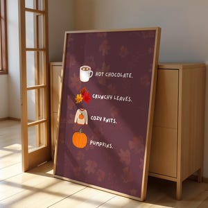 May include: A framed print with a burgundy background and autumn-themed illustrations. The text reads "Hot Chocolate," "Crunchy Leaves," "Cozy Knits," and "Pumpkins." The print is leaning against a wooden cabinet.