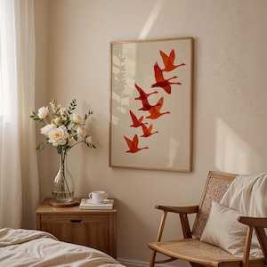 May include: A framed art print featuring a flock of orange birds in flight against a cream background. The artwork is displayed in a light-colored wooden frame. The scene is set in a bedroom with a wooden nightstand, a chair, and a vase of flowers.