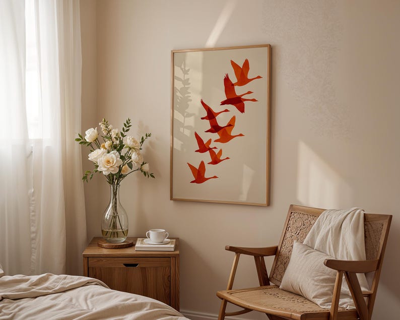 May include: A framed art print featuring a flock of orange birds in flight against a cream background. The artwork is displayed in a light-colored wooden frame. The scene is set in a bedroom with a wooden nightstand, a chair, and a vase of flowers.