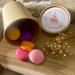 Puede incluir: Un recipiente cilíndrico de cartón marrón claro volcado de lado, derramando varias ceras aromáticas con forma de macarrón en tonos amarillo, morado, naranja, rosa intenso y rosa claro. La tapa presenta un logotipo con un cupcake y el texto Bougies Evaria.