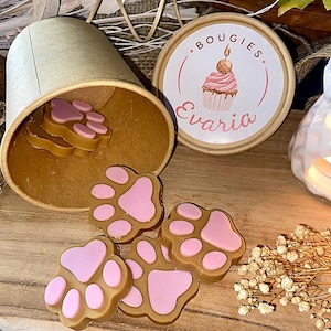 Puede incluir: Un recipiente cilíndrico de cartón con una tapa que muestra una ilustración de un cupcake y el texto "BOUGIES Evaria". Varias ceras aromáticas en forma de huella, de color beige y rosa, están esparcidas frente al recipiente abierto.