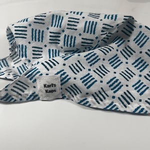 Può includere: Un copricapo in tessuto bianco caratterizzato da un motivo ripetitivo di linee tratteggiate blu ottanio scuro e piccole forme a diamante. Una piccola etichetta rettangolare in tessuto bianco è cucita sul bordo, con il testo nero Karl's Kaps.