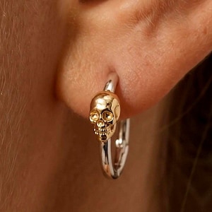 May include: A small hoop earring featuring a polished silver-tone band and a central gold-tone skull charm. The skull is detailed with defined eye sockets, a nasal cavity, and teeth, positioned directly on the front of the circular hoop earring.