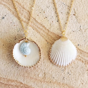 May include: Two gold chain necklaces with seashell pendants. One pendant is a white seashell with a gold rim and a light blue spiral bead. The other pendant is a white seashell with a gold rim.