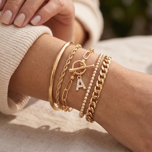 May include: A set of five gold-toned bracelets including a smooth bangle, a twisted rope chain, a paperclip chain with a toggle clasp, a beaded chain, and a curb chain. A small letter A charm with embedded clear stones hangs from the toggle.