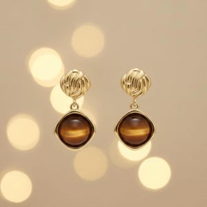 May include: A pair of gold-colored earrings. Each earring features a textured, knotted top, a small connecting loop, and a square-shaped, brown gemstone pendant. The background is a soft, blurred beige with bokeh lights.