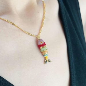 May include: A gold-colored necklace with a colorful fish pendant. The fish pendant features a variety of colors, including red, green, yellow, and blue. The fish has a gold-colored tail and is suspended from a delicate gold chain.