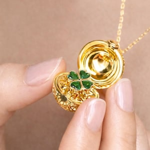 May include: A gold-colored locket necklace with a four-leaf clover design. The locket is open, revealing a green clover. The necklace is on a gold chain.