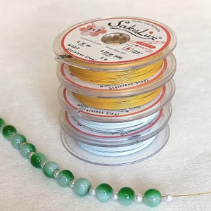 May include: A stack of four spools of beading wire in gold and white, with the brand name "SakuLux" visible. The wire is labeled as stainless steel. A strand of green and white beads is in the foreground, suggesting jewelry making.