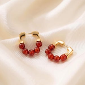 May include: A pair of gold-toned hoop earrings featuring a U-shaped metal frame with five smooth, round, deep red carnelian beads strung along the bottom curve of each earring, secured with a standard post and hinge closure mechanism.