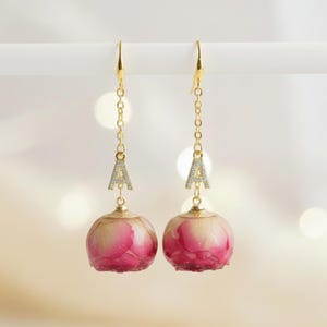 May include: A pair of dangle earrings featuring gold-colored hooks and chains. Each earring has a small, sparkling letter "A" pendant and a round, pink and white floral orb. The earrings are set against a soft, blurred background.