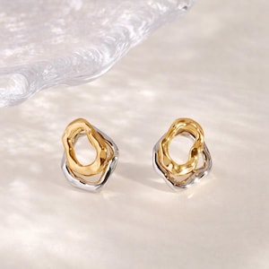 May include: A pair of modern earrings with a layered design. Each earring features a gold-colored inner loop set within a silver-colored, irregularly shaped outer frame. The earrings are displayed on a light-colored surface.