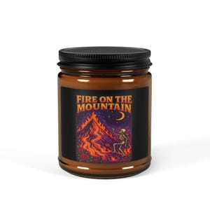 May include: A candle jar with a black lid and a label that reads "FIRE ON THE MOUNTAIN." The label features a mountain engulfed in flames, a skeleton, roses, a crescent moon, and stars. The jar is filled with a light brown wax.