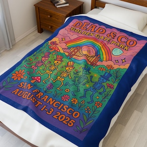 以下が含まれることがあります： 骸骨、花、虹のデザインと「DEAD & CO GOLDEN GATE PARK SAN FRANCISCO AUGUST 1-3 2025」の文字が特徴的なカラフルなブランケット。 ブランケットは青い縁取りで、ベッドの上に置かれています。