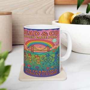 May include: White ceramic mug featuring a colorful illustration with the text "DEAD & CO GOLDEN GATE PARK" above a rainbow and the Golden Gate Bridge. Below, the text reads "SAN FRANCISCO AUGUST 1-3 2025". The design includes dancing skeletons.