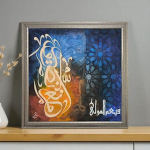 May include: Square framed artwork featuring Arabic calligraphy in cream against a vibrant background of blue, orange, and dark blue hues. The artwork is encased in a silver frame and rests on a wooden surface.