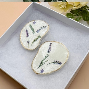 May include: Two off-white, irregularly shaped decorative pieces with gold edges, each featuring lavender sprigs. The pieces are presented in a white box with a soft, textured interior, suggesting a gift or display item.