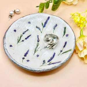 May include: A white ceramic jewelry dish with a silver rim, decorated with lavender sprigs and purple accents. A silver ring with a clear stone and a pair of pearl earrings are on the dish.