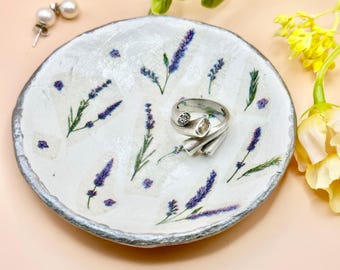 May include: A white ceramic jewelry dish with a silver rim, decorated with lavender sprigs and purple accents. A silver ring with a clear stone and a pair of pearl earrings are on the dish.