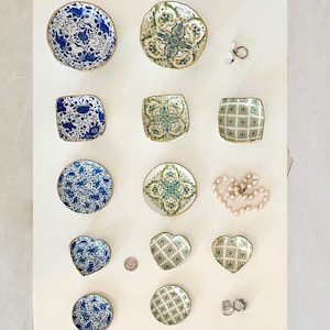 Op de afbeelding: Een verzameling decoratieve keramische schalen in verschillende vormen, waaronder rond, vierkant en hartvormig. De schalen hebben blauw-witte bloempatronen, groen-blauwe geometrische ontwerpen en gouden randen. Een parelketting en ringen zijn ook aanwezig.
