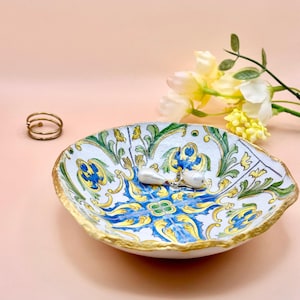 Op de afbeelding: Een decoratieve keramische schaal met een gouden rand, met een blauw, geel en groen bloemmotief. De schaal bevat een paar pareloorbellen en een gouden ring. De achtergrond is zachtroze.