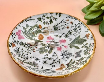 Könnte beinhalten: Eine runde weiße Schmuckschale mit goldfarbenem Rand, verziert mit einem botanischen Muster aus grünen Blättern, kleinen rosa und orangefarbenen Blüten sowie Schmetterlingen. Zwei kleine weiße Perlenohrstecker liegen in der flachen Schale.