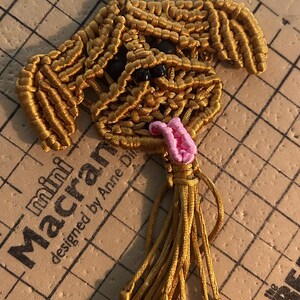May include: A golden-brown macrame dog keychain featuring a silver metal lobster clasp, dark round bead eyes, and a small pink knotted tongue. The dog shape is created with intricate cord knots and includes a long, dangling cord tassel at the base.