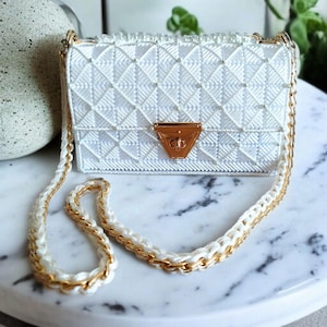 Puede incluir: Un bolso blanco tejido con un patrón geométrico. El bolso presenta un cierre dorado y una correa de cadena con detalles dorados. El bolso está sobre una superficie de mármol.