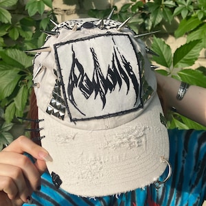 May include: A distressed, off-white baseball cap with a black and white pattern on the top. The cap features silver spikes, studs, and a black and white patch with a flame design. The brim has a silver hoop and is distressed.