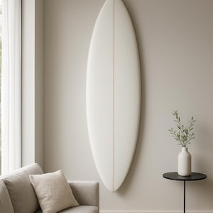 Peut inclure: Une planche de surf blanche est fixée au mur. Elle a une forme profilée avec une fine ligne centrale. Une petite table d'appoint noire avec un vase blanc et de la verdure est à proximité. Un canapé et un coussin de couleur claire sont au premier plan.