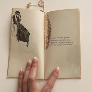 Peut inclure: Un marque-page en forme de plume dorée est inséré dans un livre ouvert. Le marque-page est orné d'une chaîne et d'un petit charme circulaire. Les pages du livre sont vieillies avec du texte. Une illustration en noir et blanc d'une femme est sur la page de gauche.