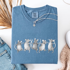 Può includere: Una maglietta blu Comfort Colors con una grafica di cinque topi grigi che si tengono per mano. Una collana d'argento con un ciondolo a forma di sole è appoggiata sulla maglietta. La maglietta è piegata, con un'etichetta visibile allo scollo.