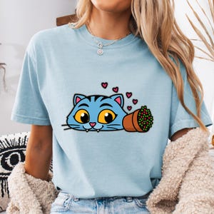 May include: Light blue t-shirt featuring a cartoon cat with blue stripes, large yellow eyes, and pink ears. The cat is next to a brown flower pot with red and green flowers, and several pink hearts. The shirt is a casual style.