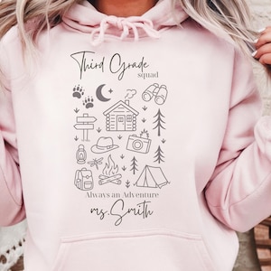 May include: A pale pink hoodie with a graphic design. The design includes the words "Third Grade squad" and "Always an Adventure", along with camping-themed illustrations like a cabin, tent, and campfire, and the name "ms. Smith."