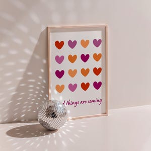 以下が含まれることがあります： 白い背景に16個のピンク、オレンジ、赤のハートがグリッド状に並んだ額装されたプリント。プリントの下部にはピンク色の筆記体で「Good things are coming」というテキストが書かれている。