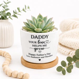 May include: A white ceramic planter containing a green succulent, resting on a round wooden base. The planter features black text that reads: DADDY YOUR love HELPS ME grow Happy Father's Day Love, Saulomyn and Theodore, with small heart icons.