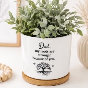 May include: A white ceramic cylindrical planter with a bamboo base containing a green leafy plant. The front features black text that reads: Dad. my roots are stronger because of you. Below the text is a black graphic of a tree with roots.