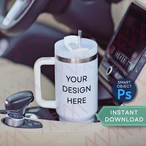 May include: White travel mug with a handle, silver trim, and a straw. The mug has the text "YOUR DESIGN HERE" printed on it. The mug is in a car's cup holder.