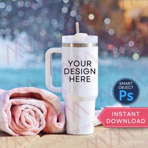 May include: A white tumbler with a handle and a straw, featuring the text "YOUR DESIGN HERE." A rolled-up pink towel rests beside it. The background shows a blurred view of water and bokeh lights.
