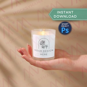 Könnte beinhalten: Eine brennende Kerze in einem transparenten Glas mit einem weißen Etikett mit einem Blumendesign und dem Text "YOUR DESIGN HERE". Die Kerze wird in einer Hand vor einem neutralen Hintergrund gehalten. Das Bild enthält auch den Text "INSTANT DOWNLOAD" und ein "SMART OBJECT"-Logo.